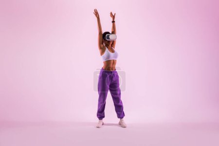 Téléchargez les photos : Exercer au-delà de la réalité. S'adapter femme noire formation dans vr casque, faire de l'exercice en métaverse, regarder tutoriel d'entraînement et échauffement sur fond de lumière néon rose, pleine longueur - en image libre de droit