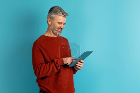 Téléchargez les photos : Joyeux bel homme d'âge moyen décontracté en utilisant une tablette numérique PC moderne sur fond bleu studio, espace de copie. Lecture d'e-book, défilement sur les médias sociaux, en utilisant l'application mobile éducative - en image libre de droit
