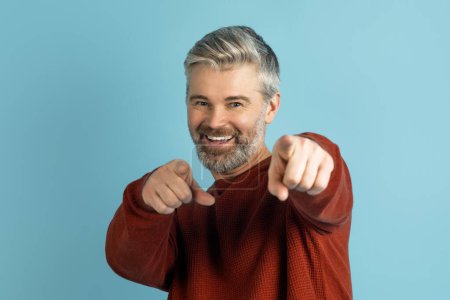 Photo for Hey you. Happy playful handsome grey-haired middle aged man in casual pointing at camera and smiling, isolated on blue studio background, flirting, enjoying good day, copy space, closeup - Royalty Free Image