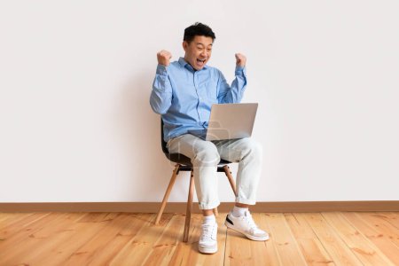 Foto de Alegre asiático de mediana edad hombre sentado en la silla con el ordenador portátil y haciendo SÍ gesto, alegre macho celebrando el éxito o el logro contra la pared de luz, sintiéndose emocionado por la gran venta o ganar en línea - Imagen libre de derechos