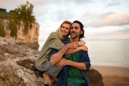 Téléchargez les photos : Portrait d'un jeune couple romantique embrassant la plage au coucher du soleil, heureux amoureux se relaxant sur les rochers près de la côte de l'océan, souriant homme et femme étreignant et regardant loin, appréciant la date, l'espace de copie - en image libre de droit