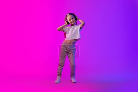 Téléchargez les photos : Petite fille d'été joyeuse et mignonne avec des tresses utilisant des écouteurs sans fil sur fond coloré lumineux, profitant d'un tout nouveau casque stéréo, écoutant sa musique préférée, espace de copie, pleine longueur - en image libre de droit