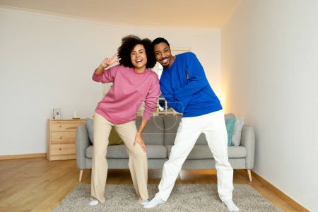 Téléchargez les photos : Joyeux noir jeune couple posant ensemble geste signe de victoire sourire à la caméra debout dans moderne salon à la maison. Pleine longueur prise de conjoints afro-américains à l'intérieur - en image libre de droit