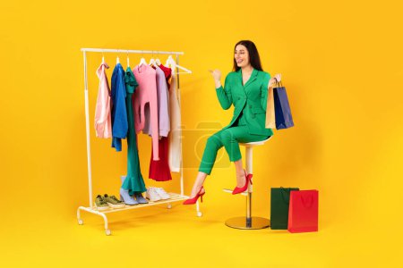Photo for Modern shopping. Happy woman shopaholic choosing clothes and pointing finger at clothing rack while buying outfit over yellow background, full length - Royalty Free Image