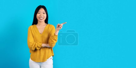Photo for Studio shot of cheerful beautiful millennial asian lady in nice outfit posing on colorful blue background, pointing at copy space and smiling, young chinese woman showing nice offer, web-banner - Royalty Free Image