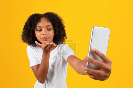 Foto de Feliz rizado adolescente afroamericano chica en camiseta blanca tomando selfie en el teléfono inteligente, soplando gesto beso aislado en el fondo del estudio amarillo. Solicitud de estudio, videollamada - Imagen libre de derechos