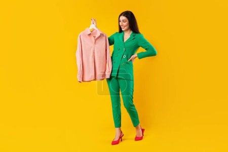 Photo for Fashion trends. Happy stylish shopaholic woman holding trendy pink jacket on hanger, standing over yellow background after successful shopping in mall, full length - Royalty Free Image