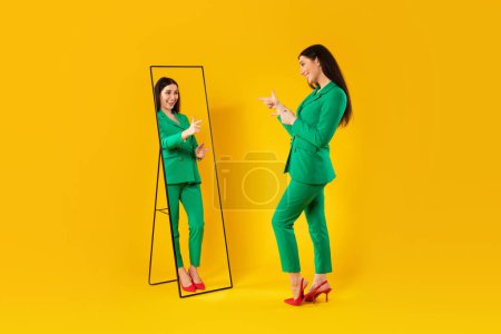 Téléchargez les photos : Dame à la mode pointant son reflet dans le miroir posant après des achats réussis, portant un costume pantalon veste verte, debout sur fond de studio jaune. J'aime ce concept de tenue - en image libre de droit