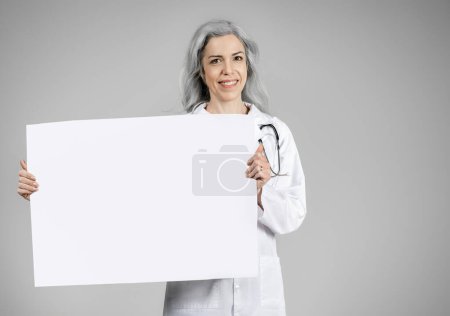Téléchargez les photos : Publicité médicale. Femme mûre médecin montrant une grande bannière en papier debout sur fond gris, portant un manteau blanc. Heureux médecin spécialiste offre de texte publicitaire. Maquette, espace de copie - en image libre de droit