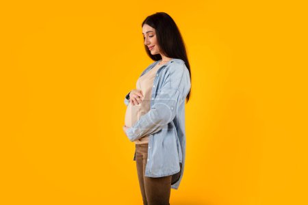 Foto de Tiempo prenatal. Joven embarazada abrazando su vientre y sonriendo, cariñosa barriga cariñosa, de pie sobre fondo amarillo del estudio, espacio para copiar - Imagen libre de derechos