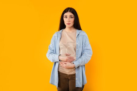 Foto de Retrato de mujer embarazada caucásica molesta tocando el vientre y mirando a la cámara, de pie sobre el fondo del estudio amarillo, espacio de copia - Imagen libre de derechos