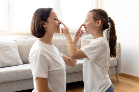 Photo for Cheerful caucasian middle aged mom and daughter in white t-shirt touch noses with hands, have fun together in living room interior. Entertainment at home, love, relationship, test of coordinate - Royalty Free Image