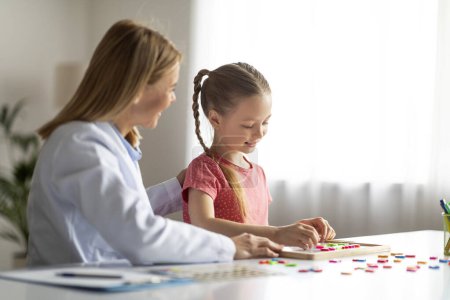 Photo for Vocabulary Building. Cute Little Girl Making Words With Colorful Letters During Therapy Session, Smiling Speech Therapist Lady Encouraging Female Child During Their Lesson In Office, Copy Space - Royalty Free Image