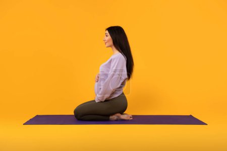 Foto de Perfil de la joven embarazada satisfecha sentada en la esterilla de fitness, practicando yoga y meditación, vista lateral, fondo amarillo. Cuidado corporal durante el embarazo, el deporte y la maternidad - Imagen libre de derechos