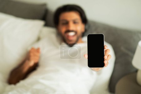 Photo for Happy millennial middle eastern guy woke up, show smartphone with blank screen, celebrating win, making success gesture, enjoy good morning in bedroom interior. Great app, ad and offer - Royalty Free Image