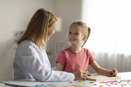 Photo for Cute Little Girl Looking At Therapist Lady During Session Meeting In Office, Smiling Female Child Sitting At Desk And Painting On Mini Chalkboard, Enjoying Development Games And Activities, Closeup - Royalty Free Image