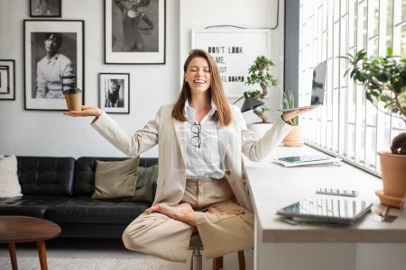 Foto de Reducir el estrés en el trabajo. Feliz joven emprendedora meditando, sentada en yoga posando brazos extendidos, sosteniendo portátil y café en las manos, trabajando en la oficina - Imagen libre de derechos