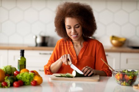 Foto de Feliz joven mujer negra cocinando ensalada de verduras frescas en la cocina, sonriendo milenaria dama afroamericana picando pepino, preparando el almuerzo en casa, disfrutando de comer alimentos saludables, copiar espacio - Imagen libre de derechos