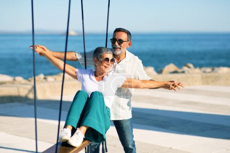 Golden Years Fun. Cheerful Mature Couple Swinging At Sea Coastline, Enjoying Sunny Summer Day Near Water Outdoors. Seniors Embracing Dream Holidays Vacation, Fooling And Laughing At Seaside