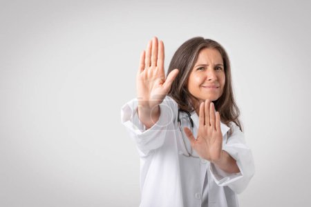 Photo for Reluctant and displeased senior woman doctor in white coat saying no, showing refusal gesture, telling to stop or stay away, standing on grey studio background, copy space - Royalty Free Image