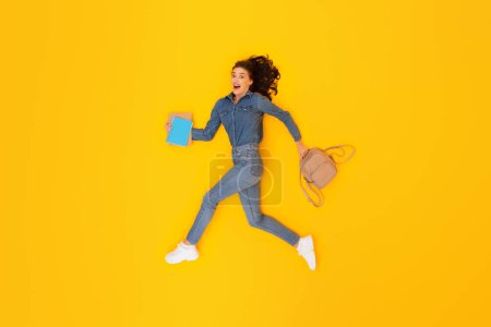 Photo for Study Enthusiasm. Emotional Lady Student With Backpack And Books In Hands Running Expressing Positivity Over Yellow Studio Background. Learner Woman Hurrying To University. Modern Education - Royalty Free Image