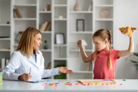 Photo for Disobedient bad tempered little girl throwing tantrum at appointment meeting with psychologist, misbehaving female child having anger issues, threatening specialist doctor with a toy, free space - Royalty Free Image
