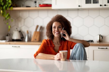 Téléchargez les photos : Joyeuse dame noire buvant du café et parlant sur un téléphone portable dans la cuisine, jeune femme afro-américaine assise à table et ayant un appel mobile, tenant une tasse et profitant d'une conversation agréable - en image libre de droit