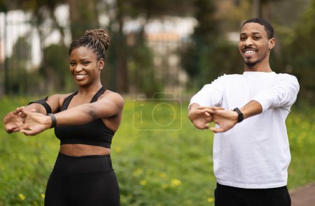Téléchargez les photos : Joyeux jeune couple noir en vêtements de sport faire échauffement et des exercices de main dans le parc d'été. Soins du corps et relation, entraînement sportif en plein air, exercice pour les muscles - en image libre de droit
