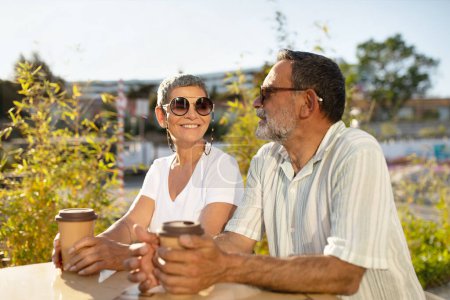 Photo for Happy Senior Couple Drinking Coffee Sitting At Outdoor Cafe On Sunny Summer Day, Holding Paper Cups With Hot Drink And Smiling To Each Other Enjoying Vacation. Retirement Lifestyle And Happiness - Royalty Free Image