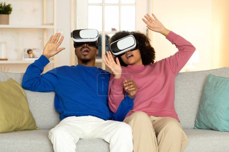 Téléchargez les photos : Aventures virtuelles. Couple Afro-Américain Incroyable S'engager dans un jeu amusant VR, portant des écouteurs assis sur le canapé à la maison. Session de jeux numériques, divertissement innovant - en image libre de droit
