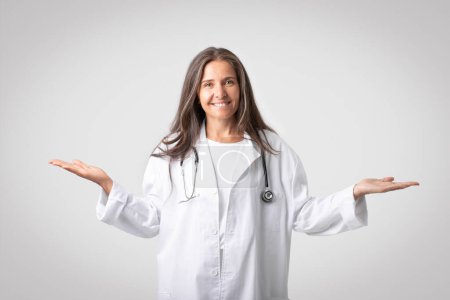 Téléchargez les photos : Femme médecin européenne joyeuse tenant quelque chose d'invisible sur les deux mains paumes souriantes à la caméra, isolée sur fond de studio gris. Mode de vie sain, santé, concept médical - en image libre de droit
