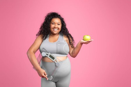 Foto de Feliz gordita dama afroamericana en ropa deportiva sosteniendo escamas y manzana verde aislada sobre fondo rosa, espacio libre. Dieta y oferta, fitness y cuidado del cuerpo, comida saludable - Imagen libre de derechos