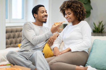 Téléchargez les photos : Aimer beau jeune homme noir mari nourrir sa petite amie, heureux couple millénaire afro-américain assis sur le canapé à la maison, manger de délicieuses pizzas ensemble, copier l'espace - en image libre de droit