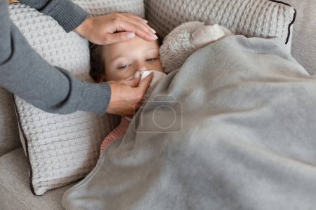 Photo for Hands of caucasian middle aged mother wipes nose in napkin to little daughter on sofa in room interior, cropped, close up. Ill care, health care, treatment and medicine at home - Royalty Free Image