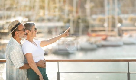 Téléchargez les photos : Offre d'été. Joyeux couple de touristes seniors en vacances pointant du doigt à l'espace libre, publicité Yacht Tour étreinte à Marina Dock en plein air. Luxe Voyage, Voilier Croisière Publicité - en image libre de droit