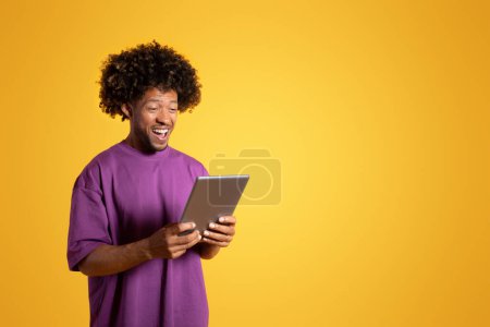 Foto de Glad inspirado sorprendido hombre rizado negro maduro en púrpura camiseta escribiendo en la tableta, aislado sobre fondo amarillo, estudio. Aplicación para comunicación, buenas noticias, redes sociales y videollamadas - Imagen libre de derechos