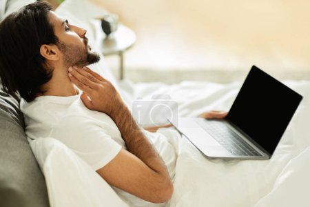 Photo for Sad middle eastern guy woke up, sits on bed, check tonsils with laptop with blank screen in bedroom interior. Sick, symptoms, health problems, medicine service and app, ad and offer - Royalty Free Image