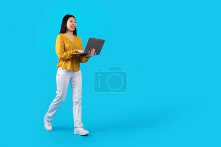 Téléchargez les photos : Jolie jeune Chinoise joyeuse en tenue décontractée étudiante utilisant un ordinateur portable moderne sur fond de studio bleu, marchant vers l'espace de copie. École en ligne, cours, e-learning, concept de webinaire - en image libre de droit