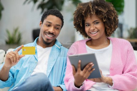 Photo for E-commerce concept. Happy smiling beautiful black couple shopping on Internet from home, sitting on couch in cozy living room, using modern digital tablet and bank card, copy space - Royalty Free Image