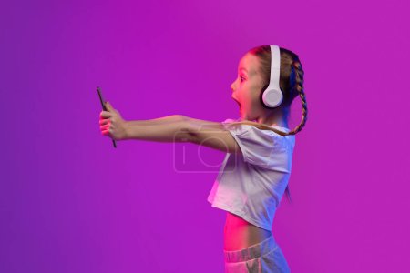 Téléchargez les photos : Émotionnel étonné petite fille preteen enfant jouer à des jeux vidéo sur tablette numérique sur fond futuriste, en utilisant des écouteurs sans fil, en regardant l'écran du pad, s'exclame. Divertissement pour enfants et gadgets - en image libre de droit