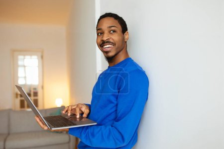 Téléchargez les photos : Technologie et travail. Happy Black Guy utilisant ordinateur portable debout près du mur à la maison. Freelance Faire du travail à distance sur l'ordinateur Surfer sur Internet et s'engager dans des loisirs en ligne. Concept d'emploi à distance - en image libre de droit