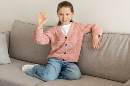 Téléchargez les photos : Joyeux caucasien petite fille agitant la main, assis sur le canapé, profiter du temps libre dans le salon intérieur. Enfance, émotions de l'enfant, salutation à l'appel vidéo, dire bonjour, salut geste à la maison - en image libre de droit