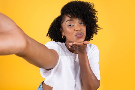Téléchargez les photos : Joyeux jeune femme frisée afro-américaine qui fait souffler baiser et selfie, amusez-vous isolé sur fond de studio jaune, gros plan. Soins de beauté, blog et médias sociaux, application photo - en image libre de droit
