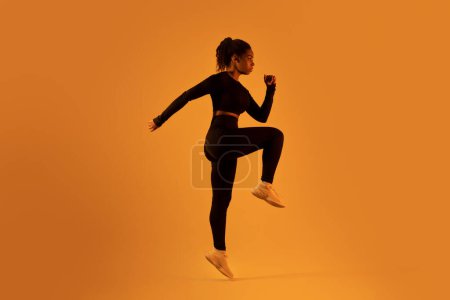 Photo for Fitness studio dedication. Determined black woman exercising, jumping and doing elbow to knee crunch over orange neon studio background, full length shot - Royalty Free Image