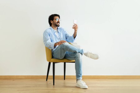 Photo for Cheerful millennial middle eastern guy blogger in glasses has video call, shooting blog on smartphone, sits on chair in room on white wall background. App and social network, communication remotely - Royalty Free Image