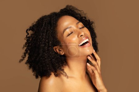 Foto de Sonriente sensual hermosa dama negra milenaria semidesnuda con el pelo tupido tocando su barbilla, fondo de estudio marrón, espacio de copia. Piel Positividad, comodidad y aceptación de la piel - Imagen libre de derechos