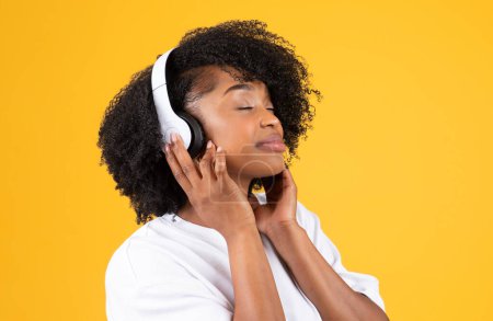 Foto de Satisfecho joven afroamericano rizado dama en auriculares inalámbricos escuchar aplicación de audio, aislado en el fondo del estudio amarillo, de cerca. Estudiante disfrutar de la música, descanso y tiempo libre - Imagen libre de derechos