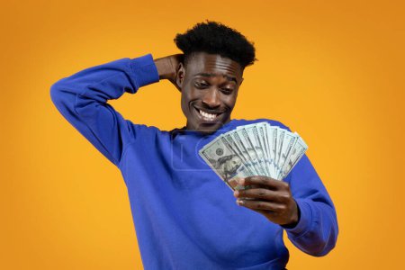Photo for Stock market investment, successful trading. Happy handsome young african american guy looking at cash dollar banknotes in his hand, touching his head and smiling on yellow studio background - Royalty Free Image