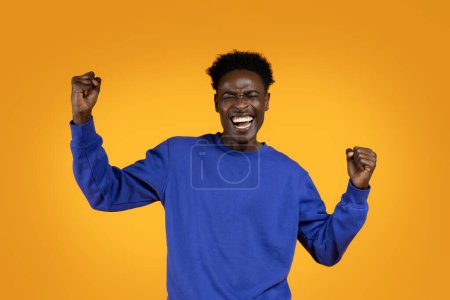 Téléchargez les photos : Émotionnel heureux heureux heureux jeune homme noir en pull bleu serrant les poings et s'exclame, isolé sur fond de studio jaune, célébrer le succès, profiter de la victoire. Émotions et gestes humains - en image libre de droit