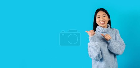 Photo for Pretty cheerful millennial asian woman in sweater pointing with thumbs at copy space on blue studio background and smiling, chinese lady recommending nice offer or deal, web-banner - Royalty Free Image